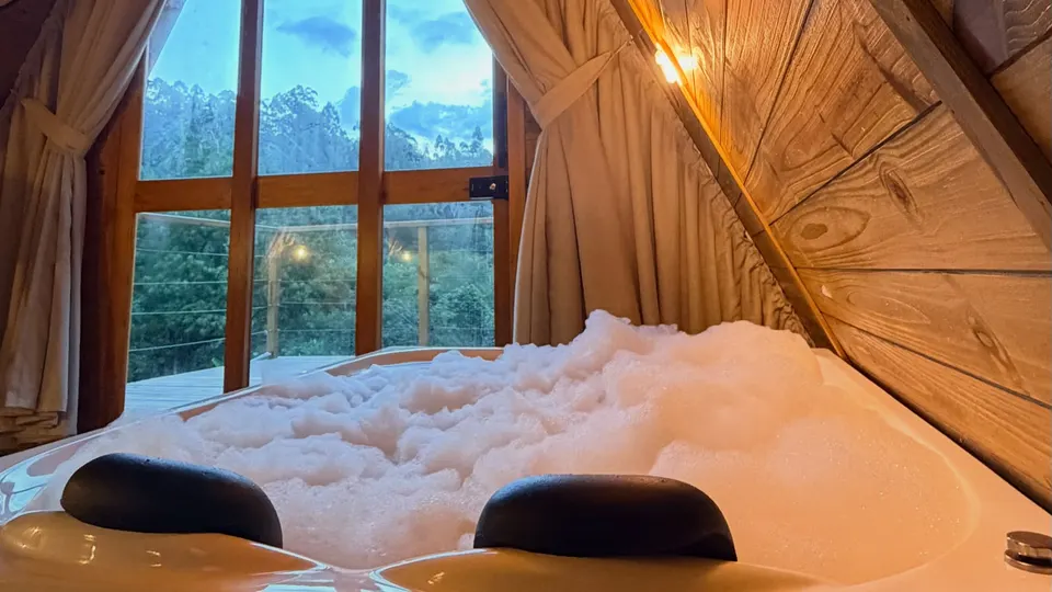 Banheira de hidromassagem com espuma diante da janela, com a mata e o céu do fim de tarde